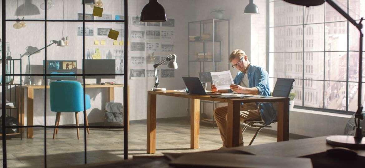 Professional Creative Designer Works on a Laptop in Loft Office, Looks at the Product Sketches and Concepts He Has Drawn. Stylish Design or Gaming Content Development Studio. Golden Hour Shot