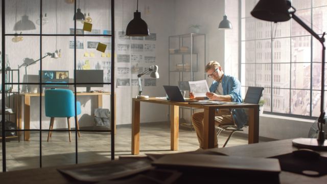 Professional Creative Designer Works on a Laptop in Loft Office, Looks at the Product Sketches and Concepts He Has Drawn. Stylish Design or Gaming Content Development Studio. Golden Hour Shot