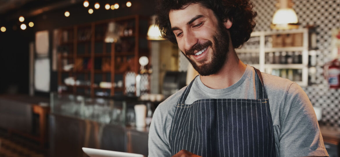 Cheerful young caucasian cafe owner wearing apron using digital tablet