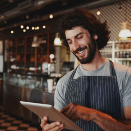 Happy young coffee shop owner holding using digital tablet