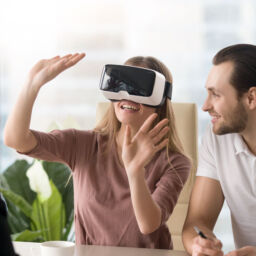 Business team of three people working on virtual reality applications and games, young excited woman testing VR glasses or goggles sitting in the office room with two colleagues, teleconference