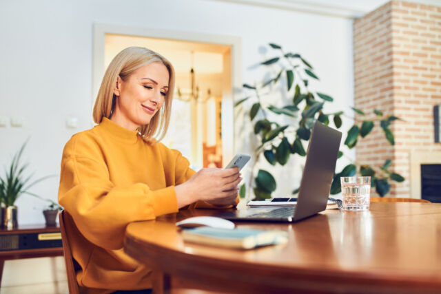 Smiling mid adult woman using smart phone while working remotely at home